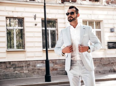 Yakışıklı, kendine güvenen, stil sahibi, lamberseksüel modelin portresi. Zarif beyaz takım elbise giymiş modern bir adam. Moda erkeği gün batımında Avrupa şehrinin arka planında poz veriyor. Güneş gözlükleriyle