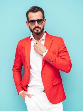 Yakışıklı, kendine güvenen, stil sahibi, lamberseksüel modelin portresi. Zarif kırmızı takım elbise giymiş modern bir adam. Moda erkeği stüdyoda mavi duvarın yanında güneş gözlüklü poz veriyor.