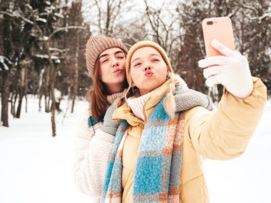 Son moda sıcak kıyafetler ve eşarplar içinde iki güzel, gülümseyen hippi kadın. Parktaki sokakta poz veren tasasız kadınlar. Pozitif saf modeller karda eğleniyor. Kış anlarının tadını çıkarıyorum. Selfie çekiyorum.