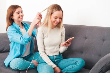 İki güzel, güzel, gülümseyen kadın. Lüks bir dairede ya da otel odasında, kanepede oturan kaygısız mankenler. Makyaj yapmak ve güzellik tedavisi yapmak. Saç düzleştirici kullanarak.