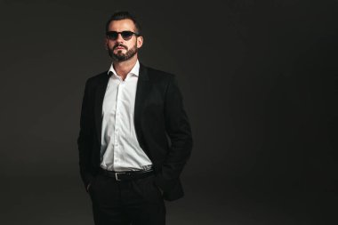 Yakışıklı, kendine güvenen, havalı hipster Lambersex modelinin yakın plan portresi. Zarif siyah takım elbiseli seksi modern adam. Moda erkeği karanlık arka planda stüdyoda poz veriyor. Güneş gözlükleriyle