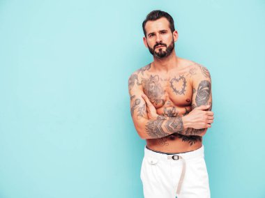 Yakışıklı, kendine güvenen, stil sahibi hipster Lambersex modelinin portresi. Seksi, modern adam. Dövmeli çıplak gövde. Moda erkeği stüdyoda açık mavi duvarın yanında poz veriyor. Çapraz kollar