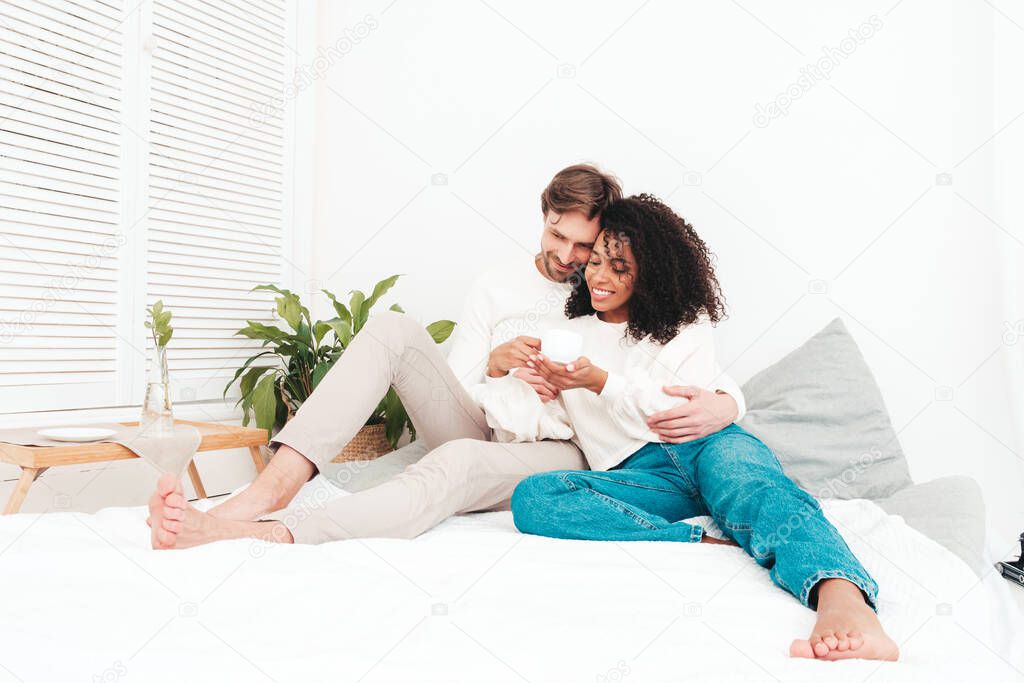 Smiling beautiful woman and her handsome boyfriend. Happy cheerful multiracial family having tender moments. Multiethnic models lying in bed and hugging in white interior. Drinking tea from cup