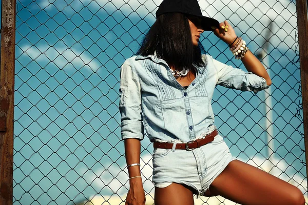 Retrato de sexy urbana moderna mujer elegante modelo de niña en pantalones vaqueros informales tela al aire libre en la calle en gorra negra detrás del cielo azul — Foto de Stock