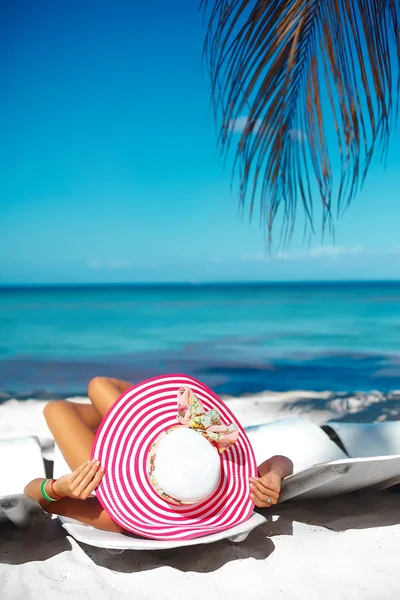 Hermosa modelo de mujer tomando el sol en la silla de playa en bikini blanco en colorido sombrero de sol detrás del océano azul de agua de verano —  Fotos de Stock