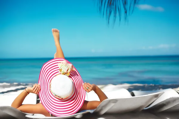 Krásná žena model opalování na pláži židli v bílých bikinách v barevné klobouku proti slunci seběhla za modré letní vody oceánu — Stock fotografie