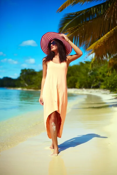 Hot beautiful woman in colorful sunhat and dress walking near beach ocean on hot summer day near palm — Stock Photo, Image