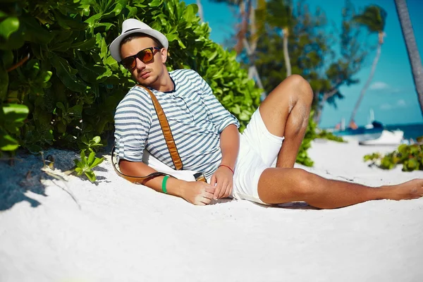 Stylish young male model man lying on beach sand  wearing hipster summer hat  enjoying summer travel holiday near ocean in sunglasses — Stock Photo, Image