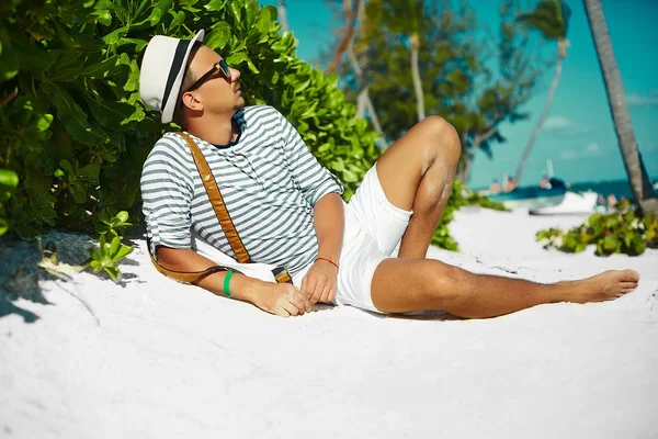 Elegante joven hombre modelo acostado en la arena de la playa con un sombrero de verano hipster disfrutando de vacaciones de verano cerca del océano en gafas de sol —  Fotos de Stock