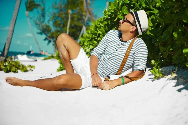 Stylish young male model man lying on beach sand  wearing hipster summer hat  enjoying summer travel holiday near ocean in sunglasses — Stock Photo, Image