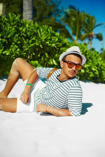 Stylish young male model man lying on beach sand  wearing hipster summer hat  enjoying summer travel holiday near ocean in sunglasses — Stock Photo, Image