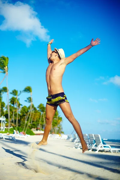 Šťastný pěkný svalnatý muž v klobouku proti slunci seběhla na pláži skákání za modrá obloha za modré oblohy — Stock fotografie