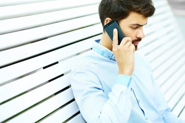 Young handsome smiling businessman model sitting on the park bench using laptop in casual hipster cloth talking on mobile phone