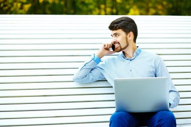 Young handsome smiling businessman model sitting on the park bench using laptop in casual hipster cloth talking on mobile phone