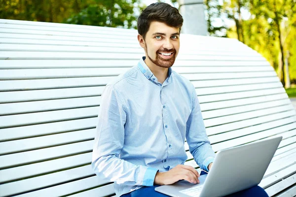 Junger gutaussehender lächelnder Geschäftsmann sitzt mit Laptop in lässigem Hipster-Tuch auf der Parkbank — Stockfoto