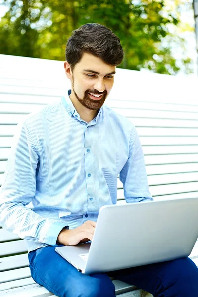 Jong knap glimlachen zakenman model zitten op het park bank met behulp van laptop in casual hipster doek — Stockfoto