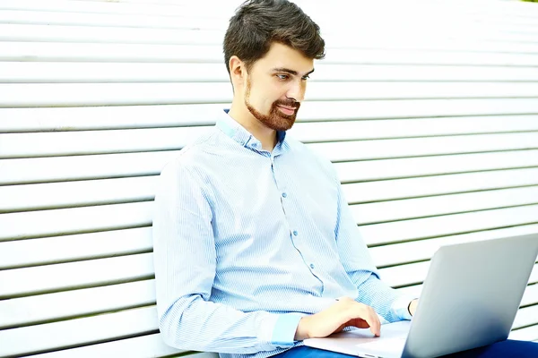 Junger gutaussehender lächelnder Geschäftsmann sitzt mit Laptop in lässigem Hipster-Tuch auf der Parkbank — Stockfoto