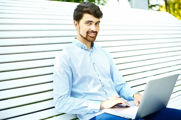 Junge hübsche lächelnde Geschäftsmann Modell sitzt auf der Parkbank mit Laptop in lässigen Hipster-Tuch reden auf dem Handy — Stockfoto