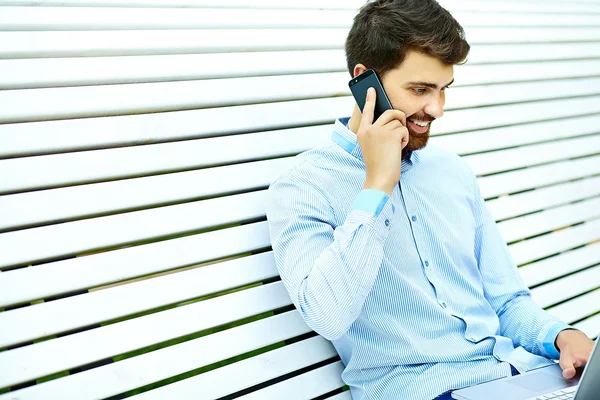 Young handsome smiling businessman model sitting on the park bench using laptop in casual hipster cloth talking on mobile phone — Stock Photo, Image