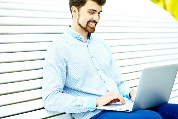 Junger gutaussehender lächelnder Geschäftsmann sitzt mit Laptop in lässigem Hipster-Tuch auf der Parkbank — Stockfoto