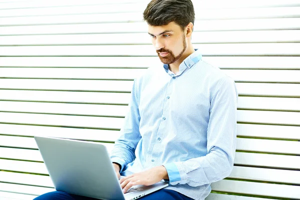 Junger gutaussehender lächelnder Geschäftsmann sitzt mit Laptop in lässigem Hipster-Tuch auf der Parkbank — Stockfoto