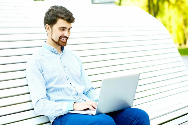 Jong knap glimlachen zakenman model zitten op het park bank met behulp van laptop in casual hipster doek — Stockfoto