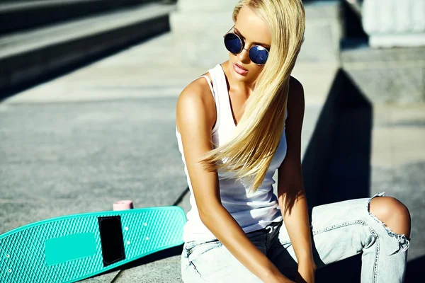 Alta moda look.glamour elegante sexy bella giovane ragazza modello biondo in estate abiti casual hipster luminosi con skateboard seduto in strada — Foto Stock