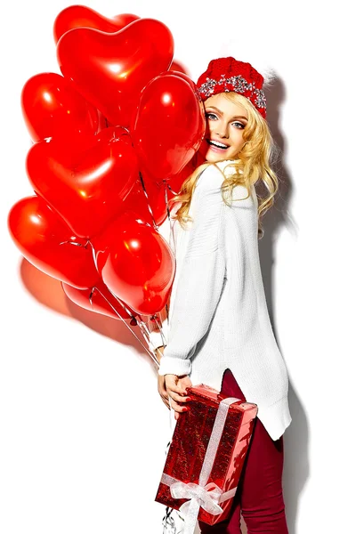 Retrato de hermosa mujer rubia sonriente dulce feliz sosteniendo en sus manos gran caja de regalo de Navidad y globos de corazón en ropa de invierno hipster rojo casual, en suéter blanco cálido —  Fotos de Stock