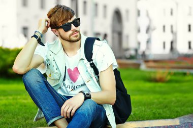 Funny smiling hipster handsome man guy in stylish summer cloth in the street sitting on grass in the park