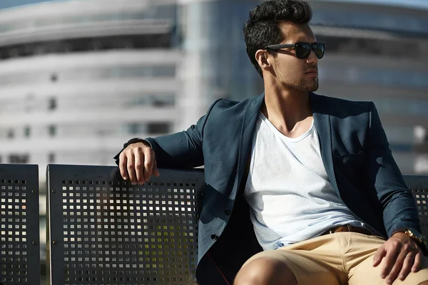 Fashion portrait of young sexy businessman handsome model man in casual cloth suit in sunglasses sitting on a bench in the street — Stock Photo, Image