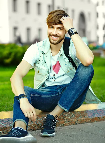 Divertido sonriente hipster guapo hombre en elegante paño de verano en la calle sentado en la hierba en el parque — Foto de Stock