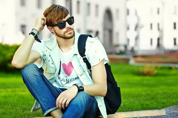 Lustig lächelnder Hipster gutaussehender Mann in stylischem Sommerkleid auf der Straße sitzend auf Gras im Park — Stockfoto