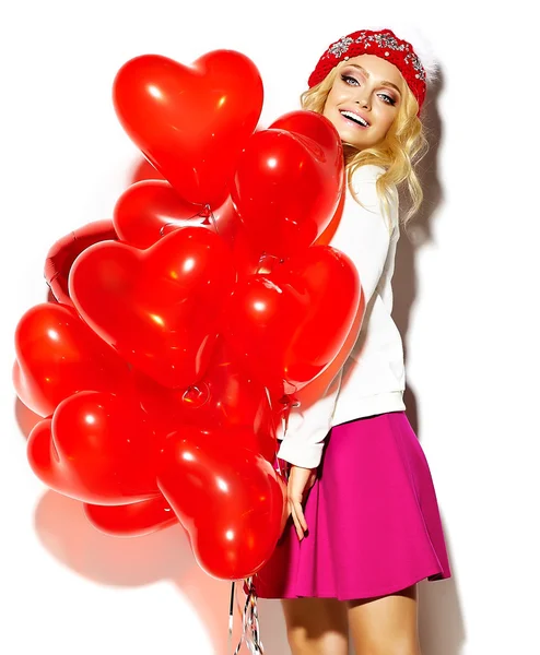 Retrato de hermosa feliz dulce linda chica rubia sonriente en ropa hipster casual, en falda rosa y gorro caliente de invierno con globos de corazón rojo en las manos —  Fotos de Stock