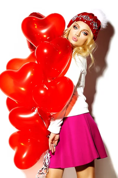 Portrait of beautiful happy sweet cute smiling blonde woman girl in casual hipster clothes, in pink skirt and winter warm beanie with red heart balloons in hands — Stock Photo, Image