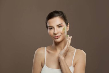 Kahverengi arka planda cilde bitkisel yüz kremi süren genç bir kadın.