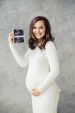 Neşeli hamile kadın ultrason görüntüsü gösteriyor ve griye gülümsüyor.