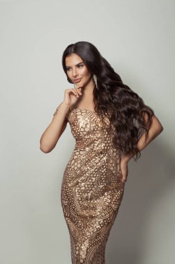 Uzun kıvırcık saçlı, beyaz arka planda altın rengi elbise giyen mutlu bir genç kadın.