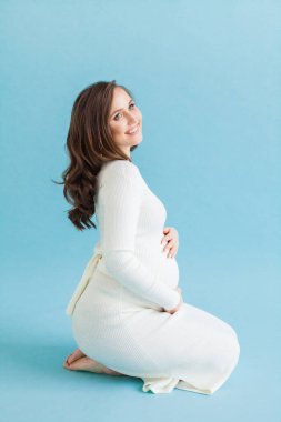 Mavi arka planda oturan beyaz elbiseli, mutlu, çekici hamile kadın.