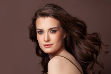 Açık tenli, esmer ve kahverengi arka planda sağlıklı kıvırcık saçlı neşeli kadın portresi.