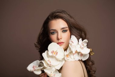 Uzun kıvırcık saçları ve kahverengi arka planda beyaz çiçekleri olan mükemmel model bir kadın.