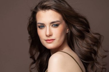 Güzel, gülümseyen bir esmer kadın portresi, açık tenli ve kahverengi arka planda sağlıklı dalgalı saçlar. Doğal makyajlı güzel bir kadın.