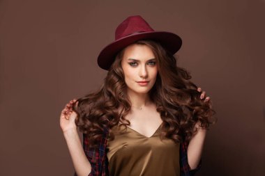 Marsala şapkası takan uzun kahverengi kıvırcık saçlı güzel bir kadının yüzü. Dalgalı saç stili olan manken. Stüdyoda poz veren kıvırcık saçlı çekici bir kadın.