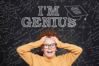 Dahi çocuk portresi eğleniyor. Mutlu kızıl saçlı çocuk kara tahtaya karşı