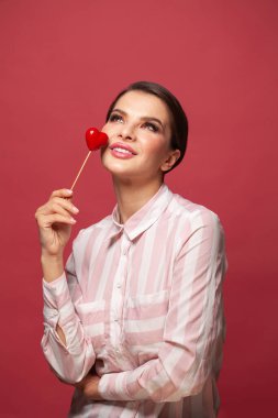 Gülümseyen, hayal kuran ve pembe arka planda kırmızı kalbi tutan genç bir kadın.