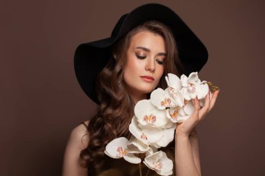 Esmer model kadın, uzun kıvırcık saçlı, siyah klasik şapka portresi takıyor.