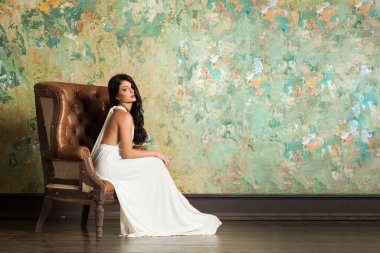 Uzun saçlı ve makyajlı zarif bir kadın lüks bir iç mekanda oturuyor.