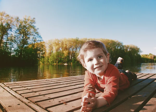 Portrait of a smiling boy laying on a dock at a sunny lake — Stock Photo, Image