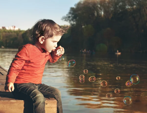 Little boy blowing soap bubbles at a lake — Stock Photo, Image