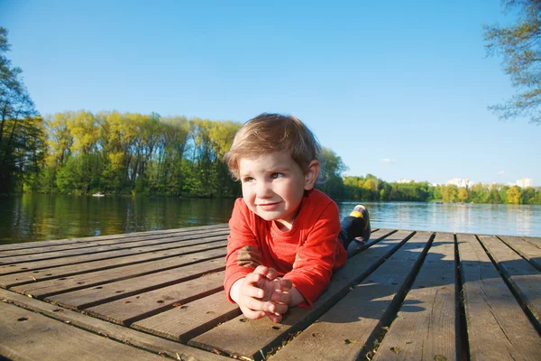 Retrato de un niño sonriente tendido en un muelle en un lago soleado —  Fotos de Stock