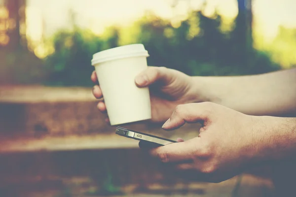 Mãos usando um telefone e segurando xícara de papel de café — Fotografia de Stock